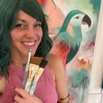 Woman with green hair smiling, holding paintbrushes, in front of colorful abstract background with a bird silhouette.