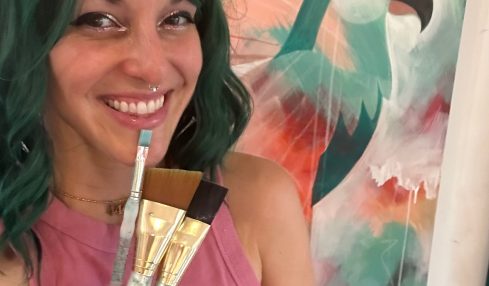 Woman with green hair smiling, holding paintbrushes, in front of colorful abstract background with a bird silhouette.