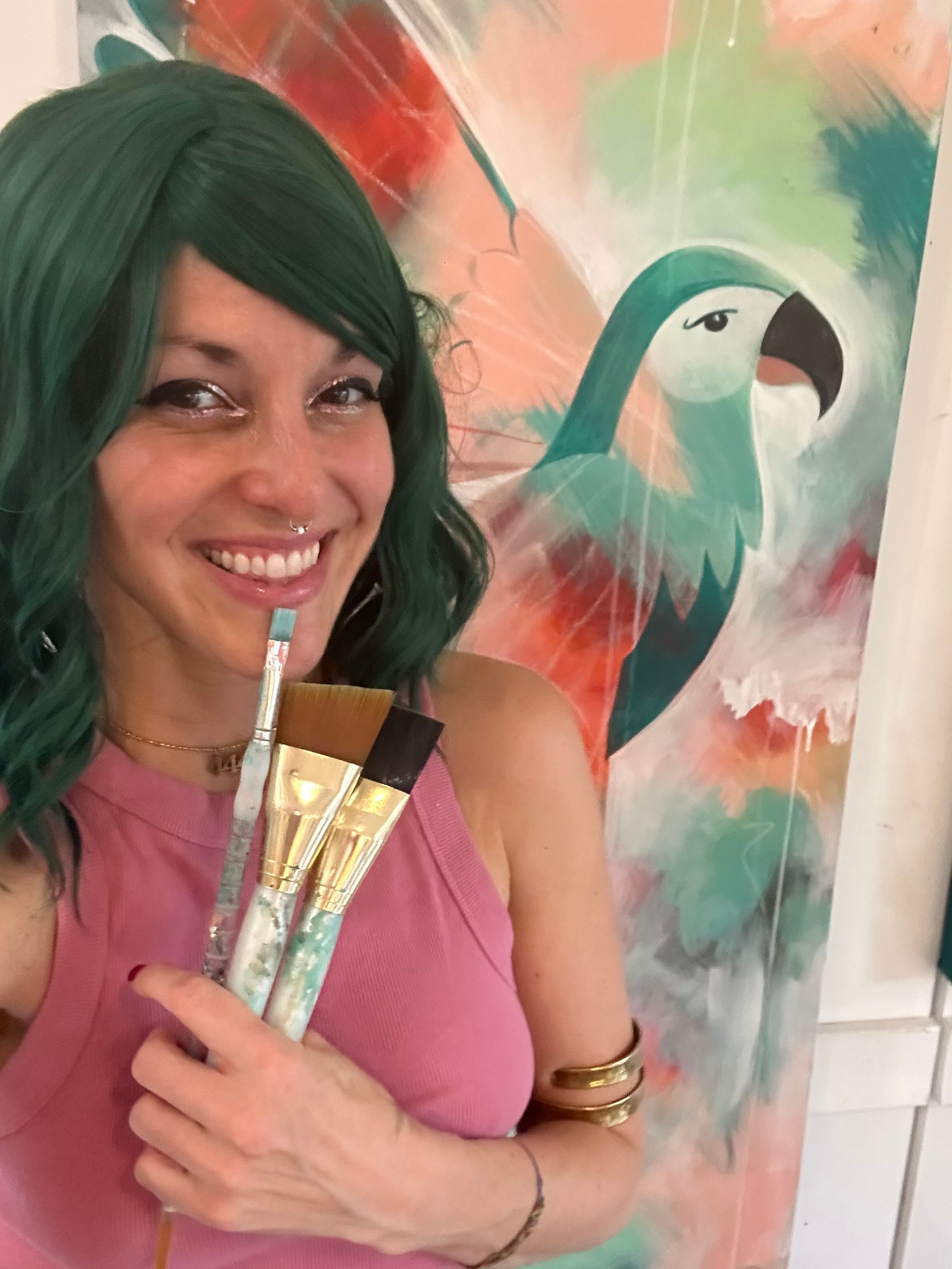 Woman with green hair smiling, holding paintbrushes, in front of colorful abstract background with a bird silhouette.