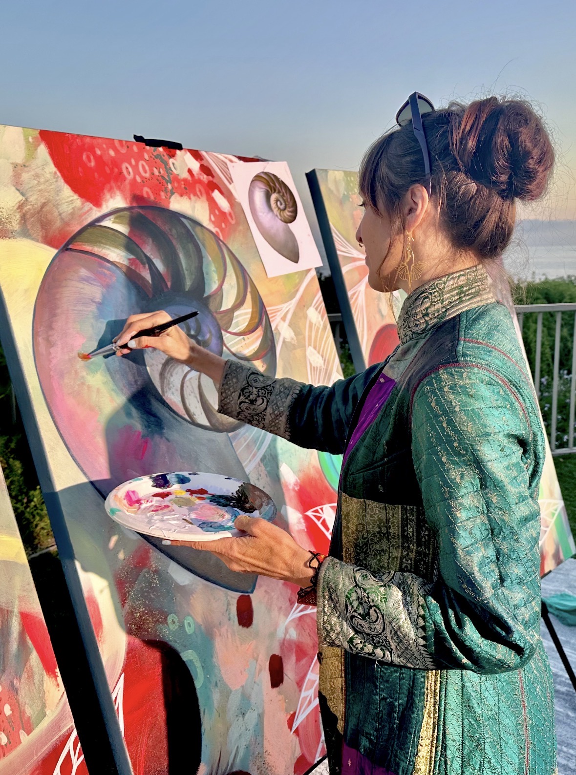 Woman painting a colorful abstract portrait on an outdoor canvas, holding a palette and brush, with a clear sky background.
