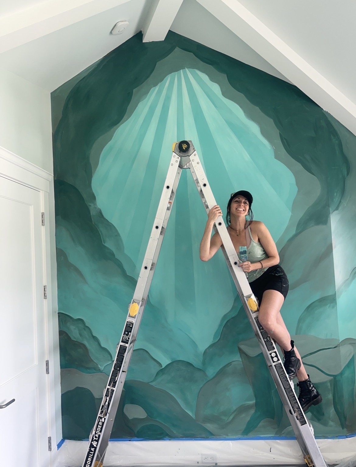 Person standing on a ladder in front of a painted mountain scene with rays of light, holding a paintbrush, smiling.