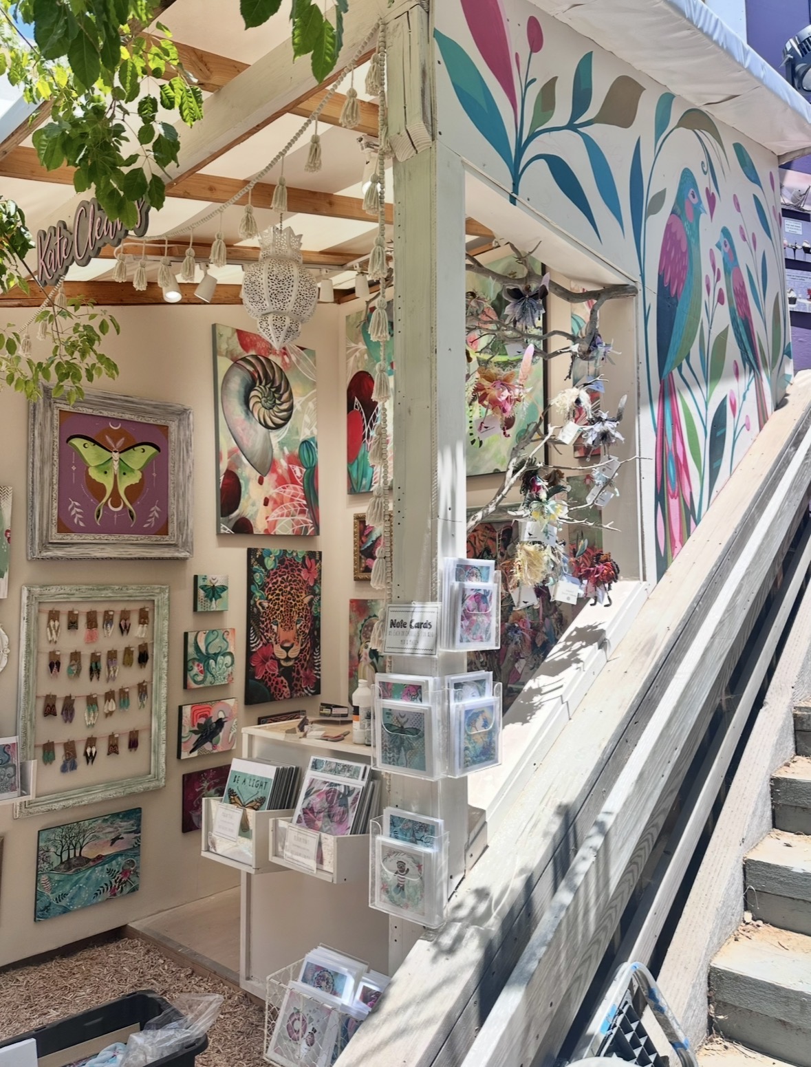 Art booth with colorful paintings, crafts, and hanging decorations inside a white tent, viewed from outside on a staircase.