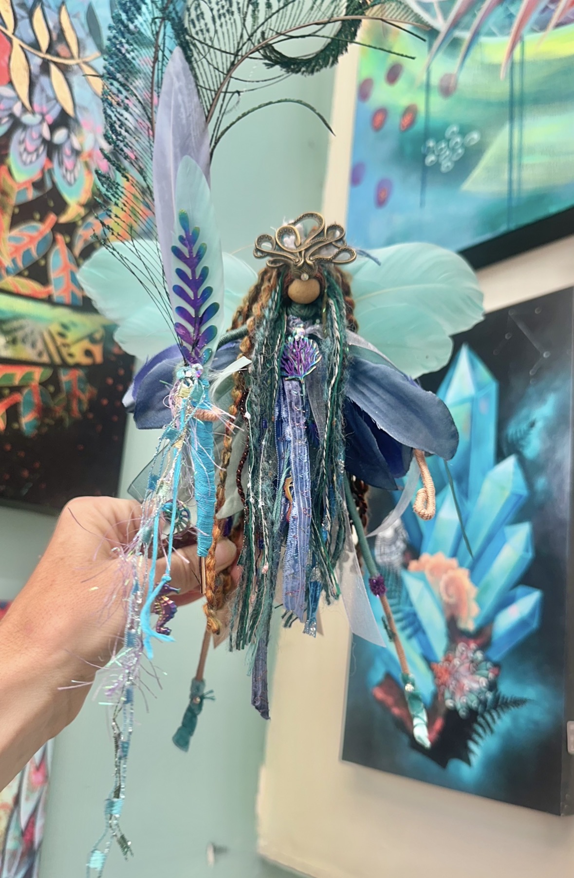 Hand holding a doll with long, colorful yarn hair, surrounded by vibrant, abstract artwork in the background.