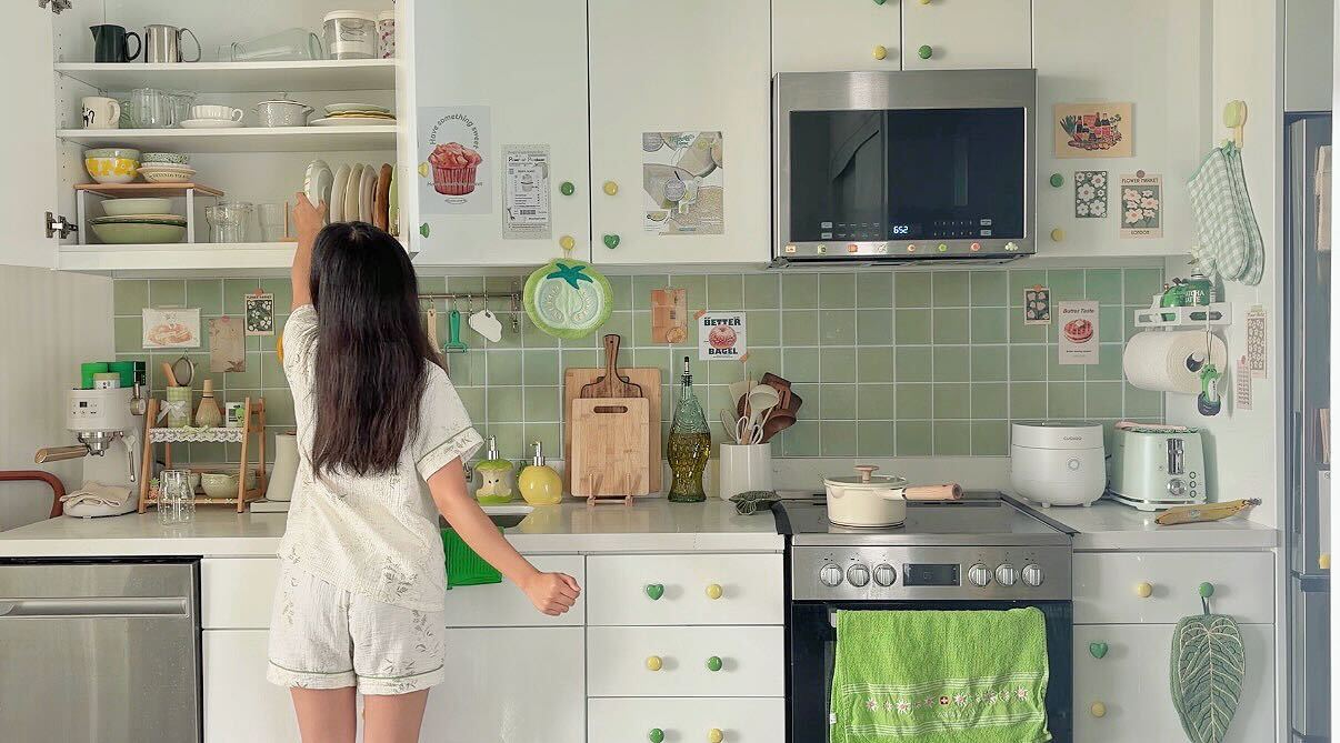 Child with long dark hair in a white dress stands in a kitchen with green tiles and white cabinets, facing away.