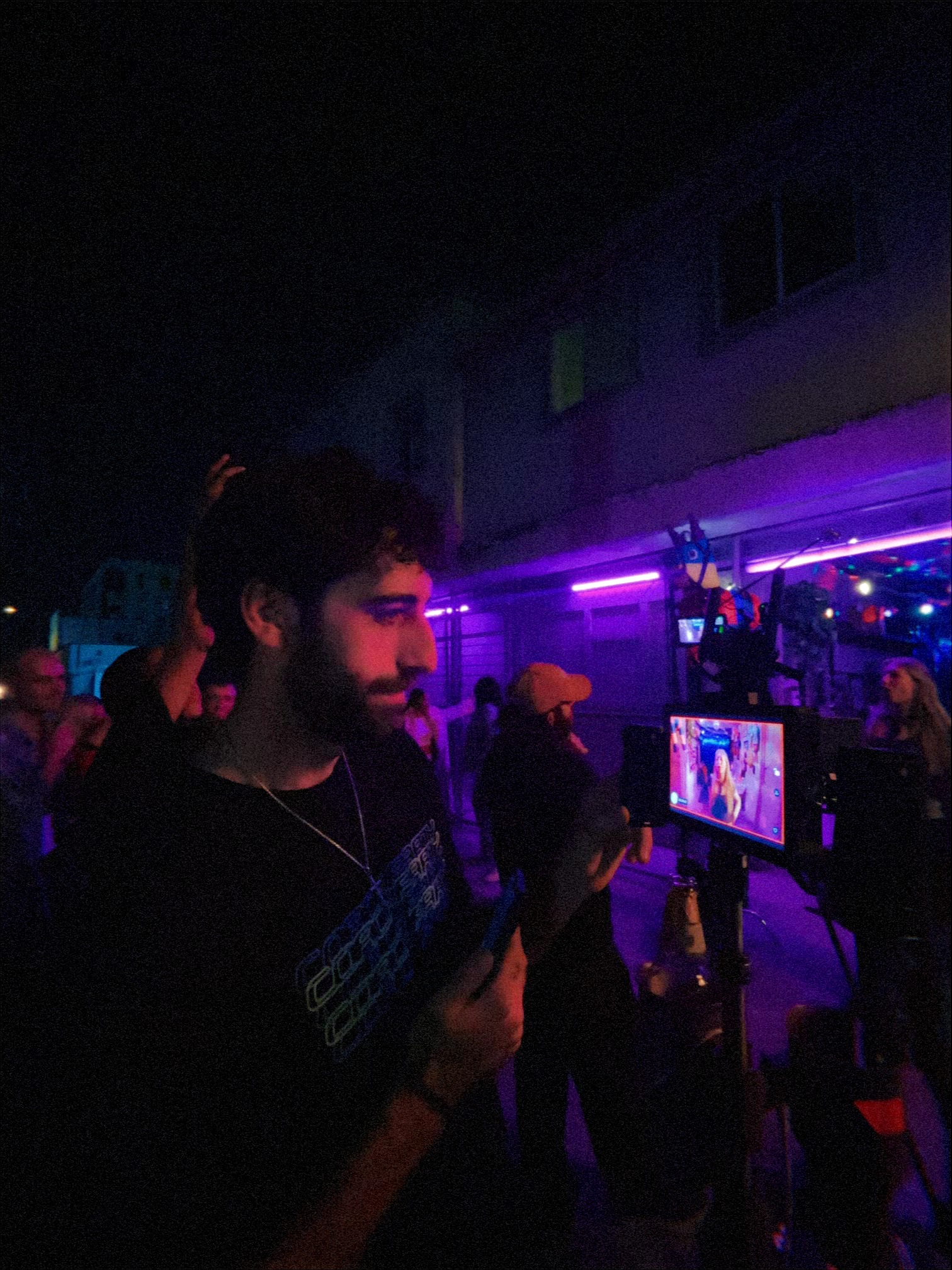 Person with beard looking at a tablet in a dark outdoor setting with purple neon lights.
