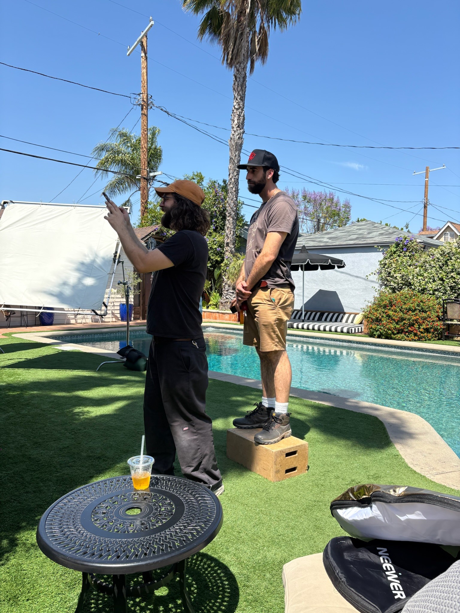 Two people standing near a swimming pool, one on a box, with a person in black and a person in gray shirt and shorts, outdoors on a sunny day.