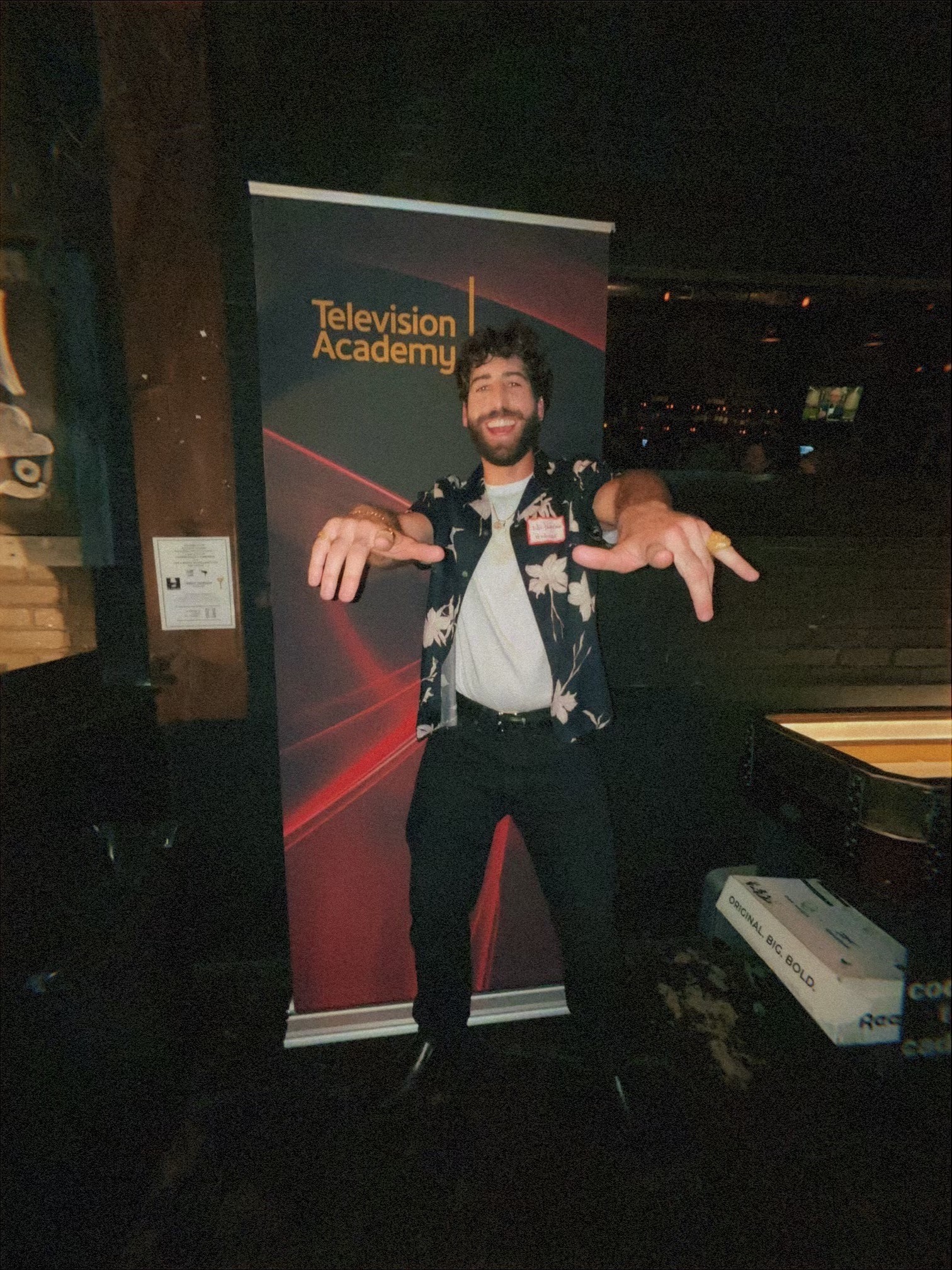 Person with beard and glasses standing in front of a TV Academy banner, smiling and gesturing with hands, in a dark room.