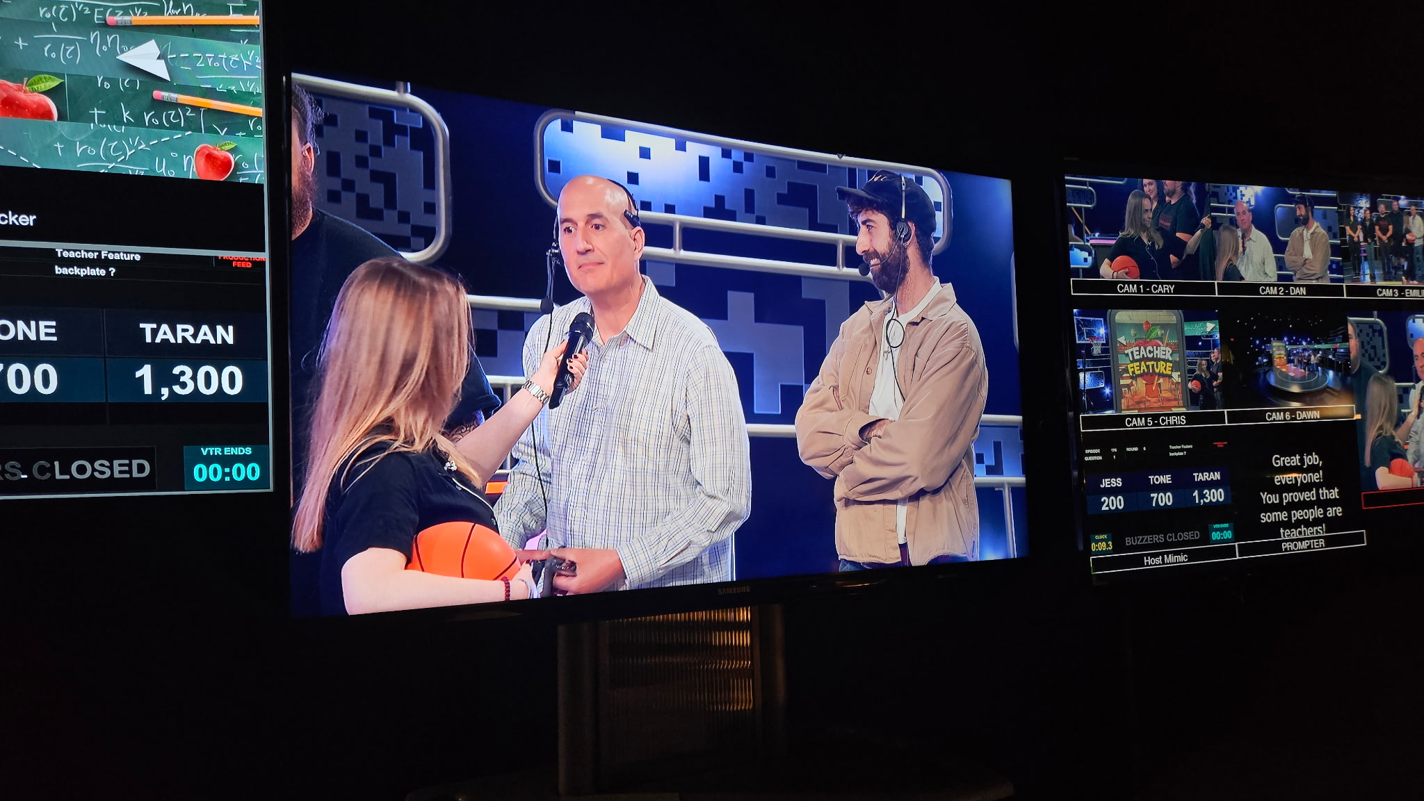 Television screen showing a woman interviewing two men in a studio with multiple screens in the background.