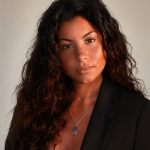 Portrait of a woman with long, curly hair wearing a black blazer and a necklace with a blue pendant.
