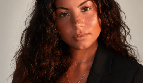 Portrait of a woman with long, curly hair wearing a black blazer and a necklace with a blue pendant.