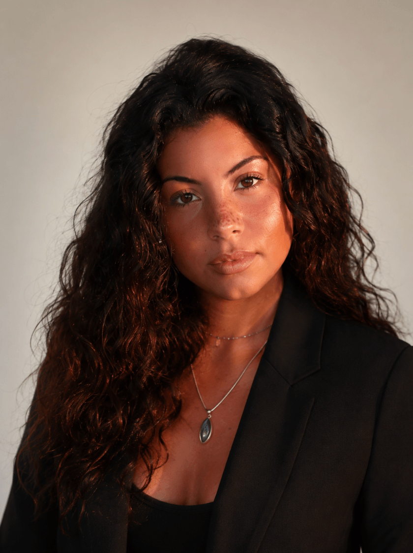 Portrait of a woman with long, curly hair wearing a black blazer and a necklace with a blue pendant.