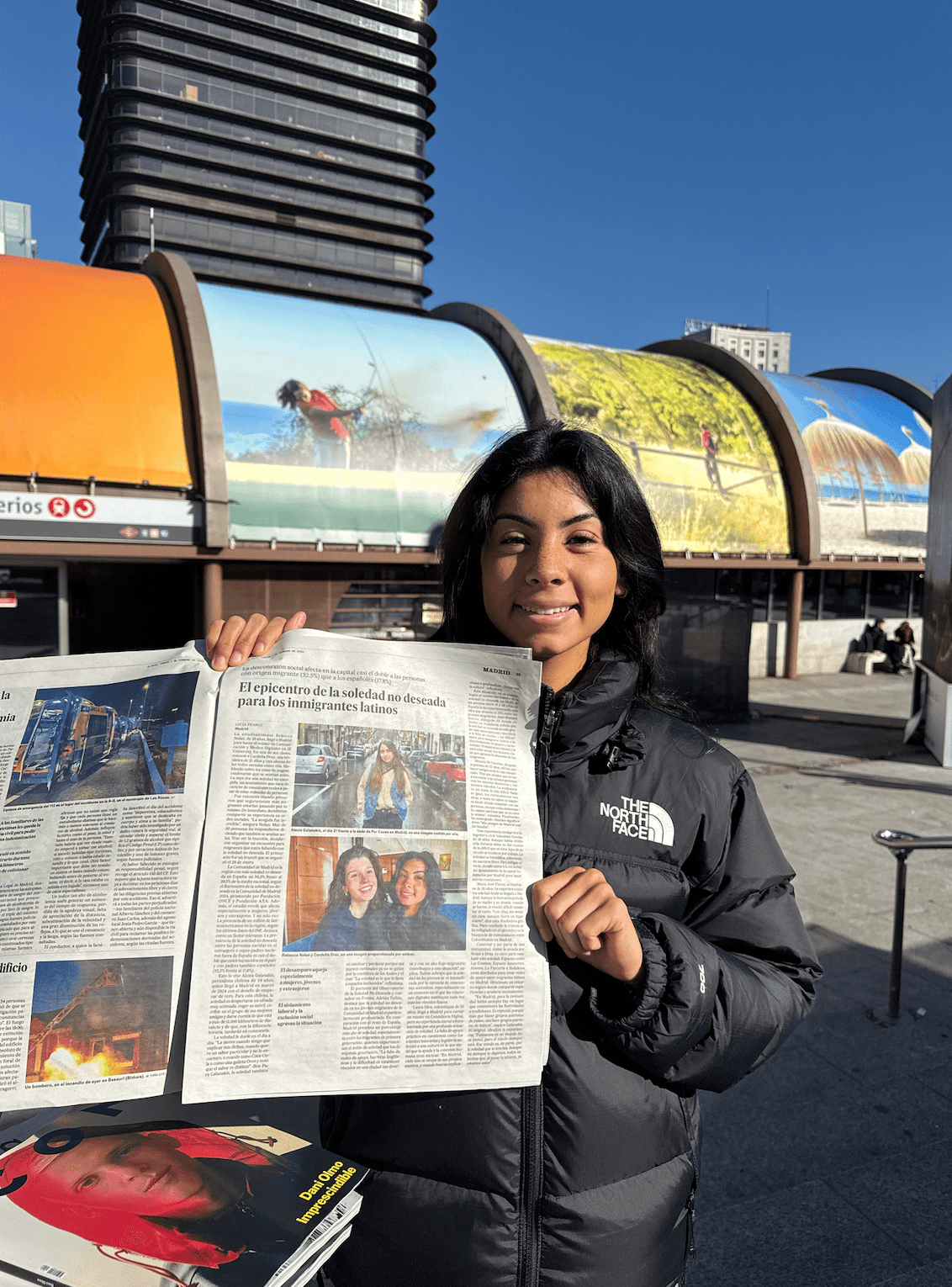 Young woman holding a newspaper outdoors with colorful structures and a building in the background.