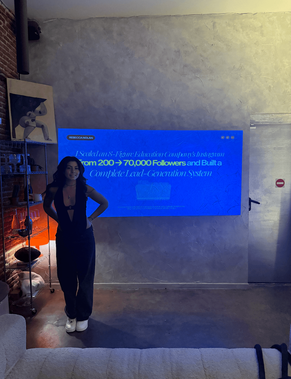 Person standing in front of a blue screen with text, in a room with a concrete wall and shelves.