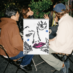 Three people sitting around a drawing board with a large sketch of a face, surrounded by greenery.