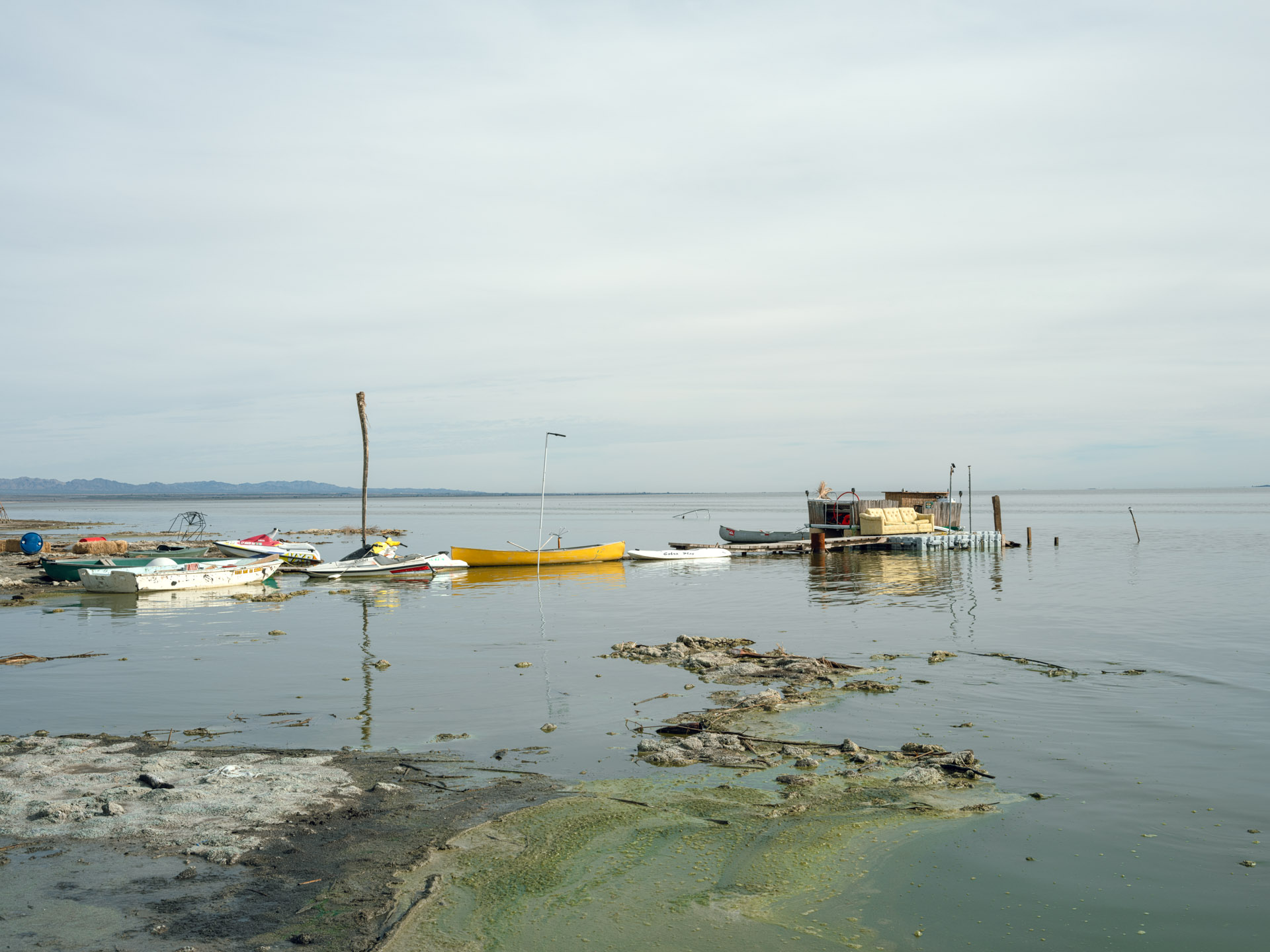 Calm water with boats and a small structure, overcast sky, distant shoreline, and some rocks in the foreground.