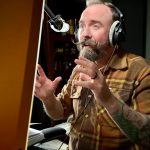 Man with a beard and tattoos wearing headphones speaking into a microphone in a recording studio.