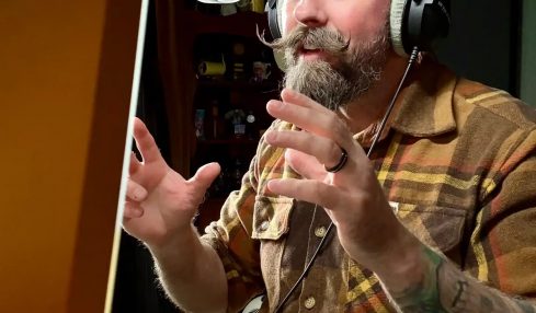 Man with a beard and tattoos wearing headphones speaking into a microphone in a recording studio.