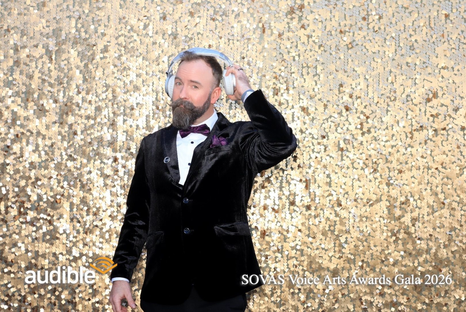Man in tuxedo with bow tie and beard, standing against a gold glitter background, touching his head with one hand.