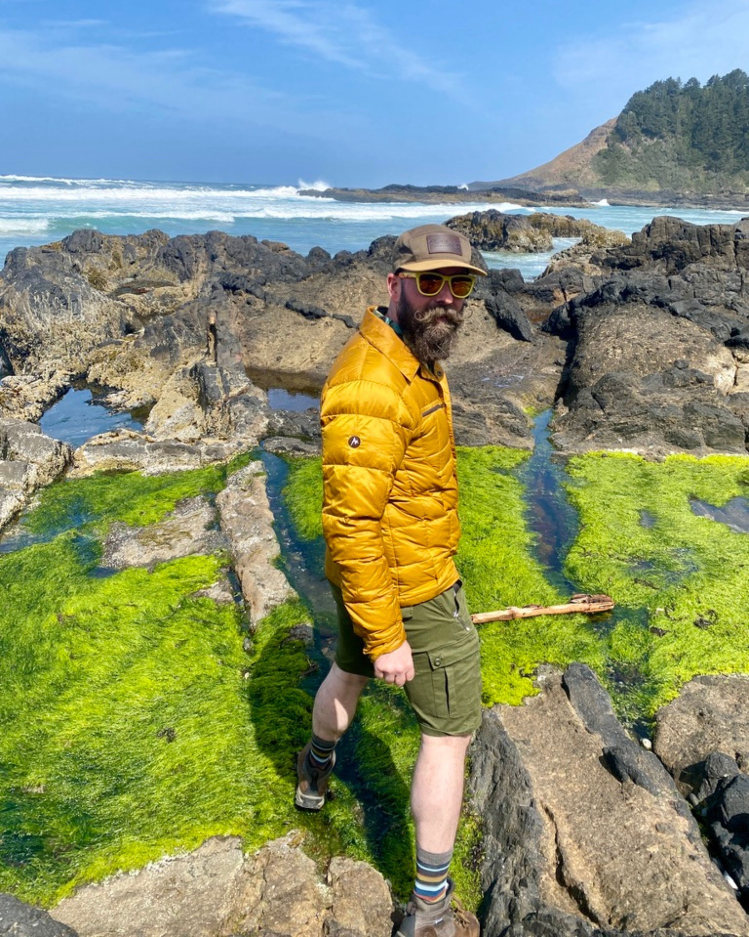 Man with a beard in a yellow jacket, sunglasses, and hat stands on rocks near the ocean with green moss and a distant shoreline.