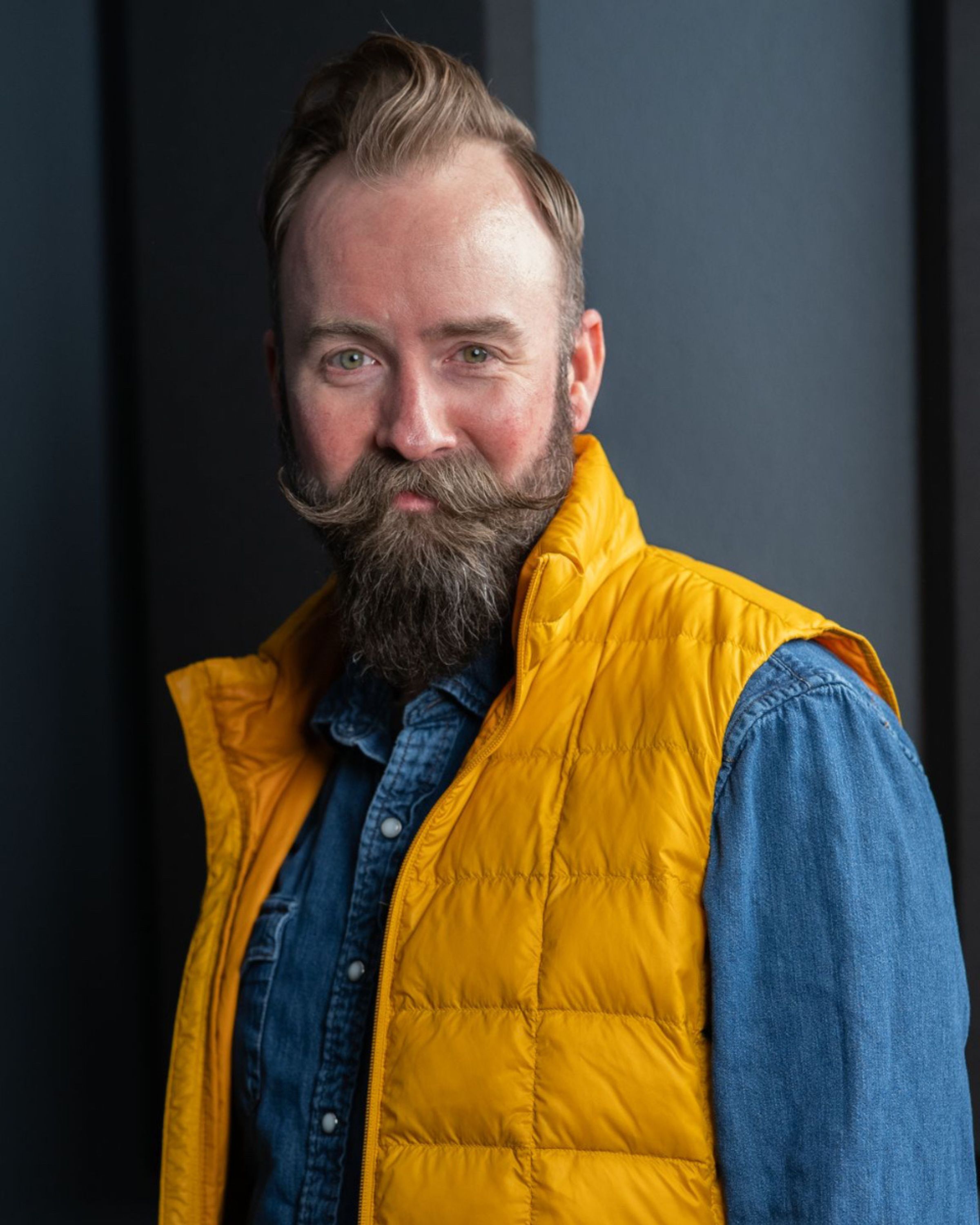 Man with a beard and mustache wearing a yellow vest over a blue shirt, standing indoors.