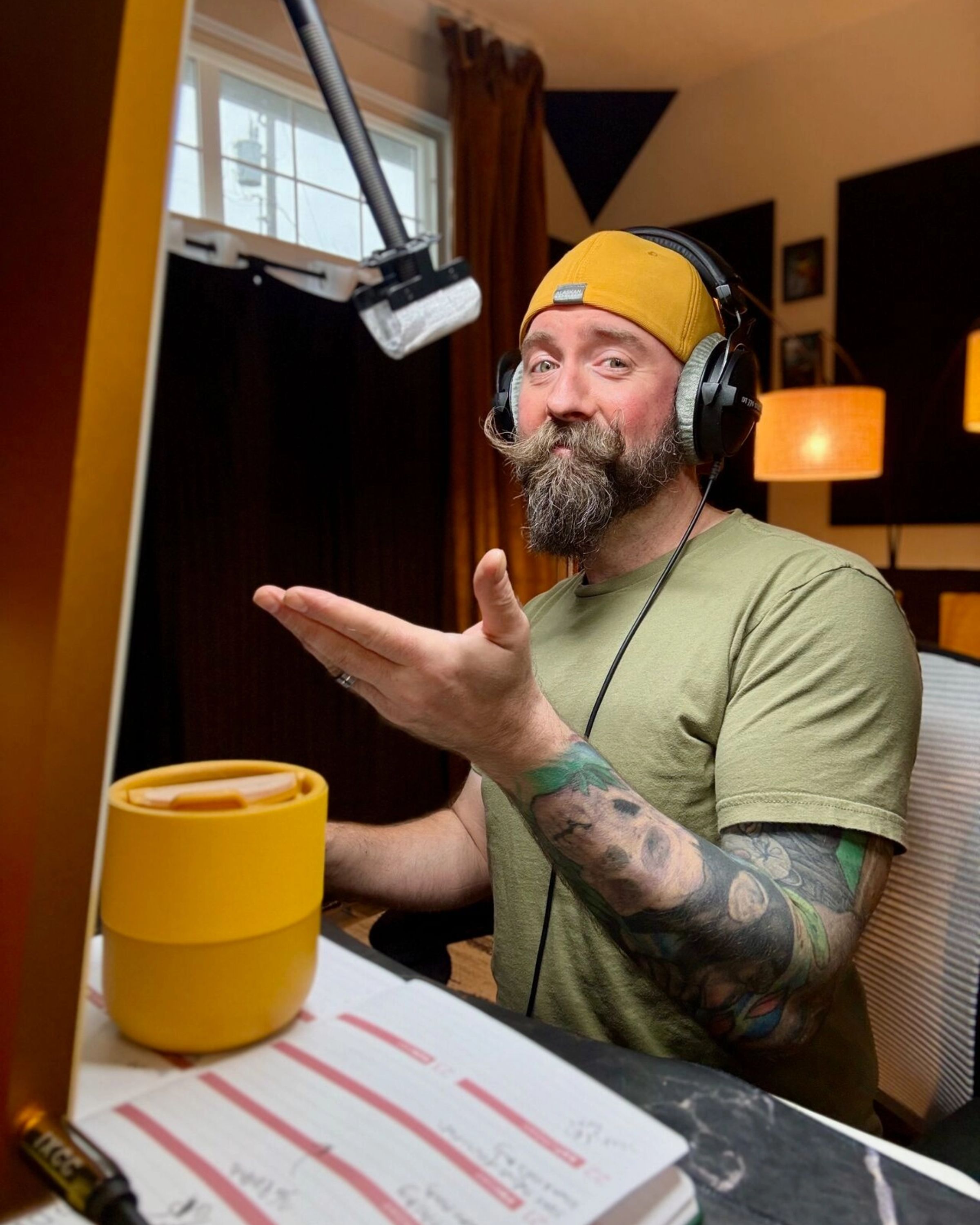 Man with a beard and tattoos wearing headphones and a yellow cap, speaking into a microphone in a recording studio.