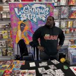 Man standing at a table with comic books, in front of a colorful graffiti-style sign reading 'Lighting Strike'.