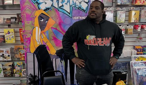 Man standing at a table with comic books, in front of a colorful graffiti-style sign reading 'Lighting Strike'.