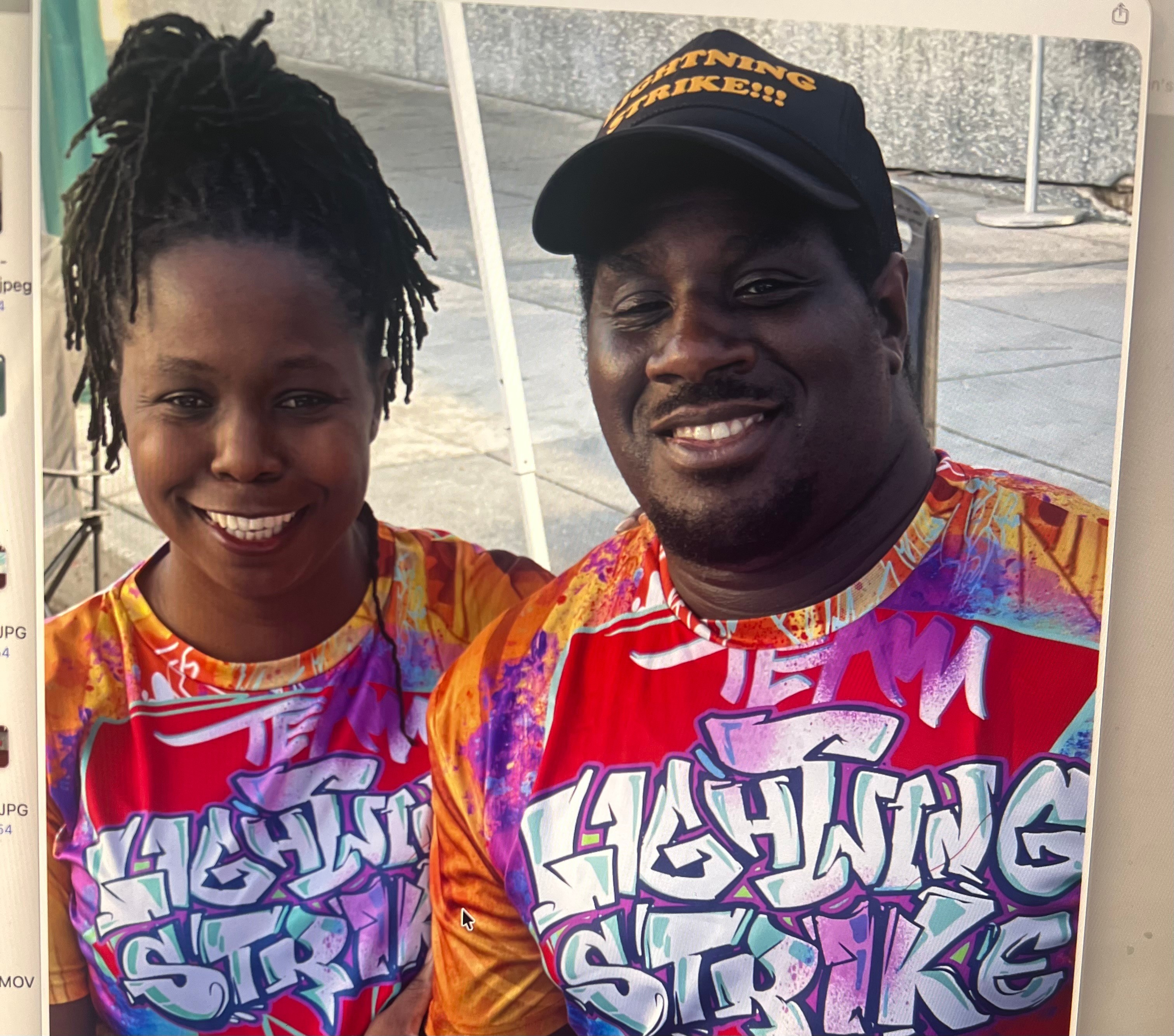 Two smiling people wearing colorful shirts with graffiti-style text, standing outdoors with a railing in the background.