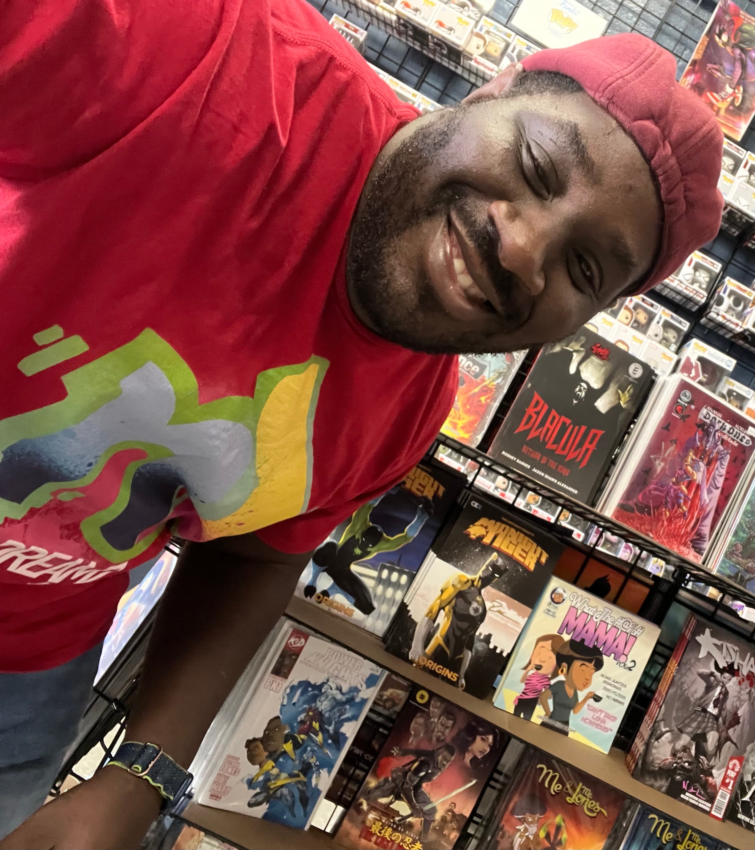 Smiling man wearing a red cap and T-shirt in a store with comic books and collectibles behind him.