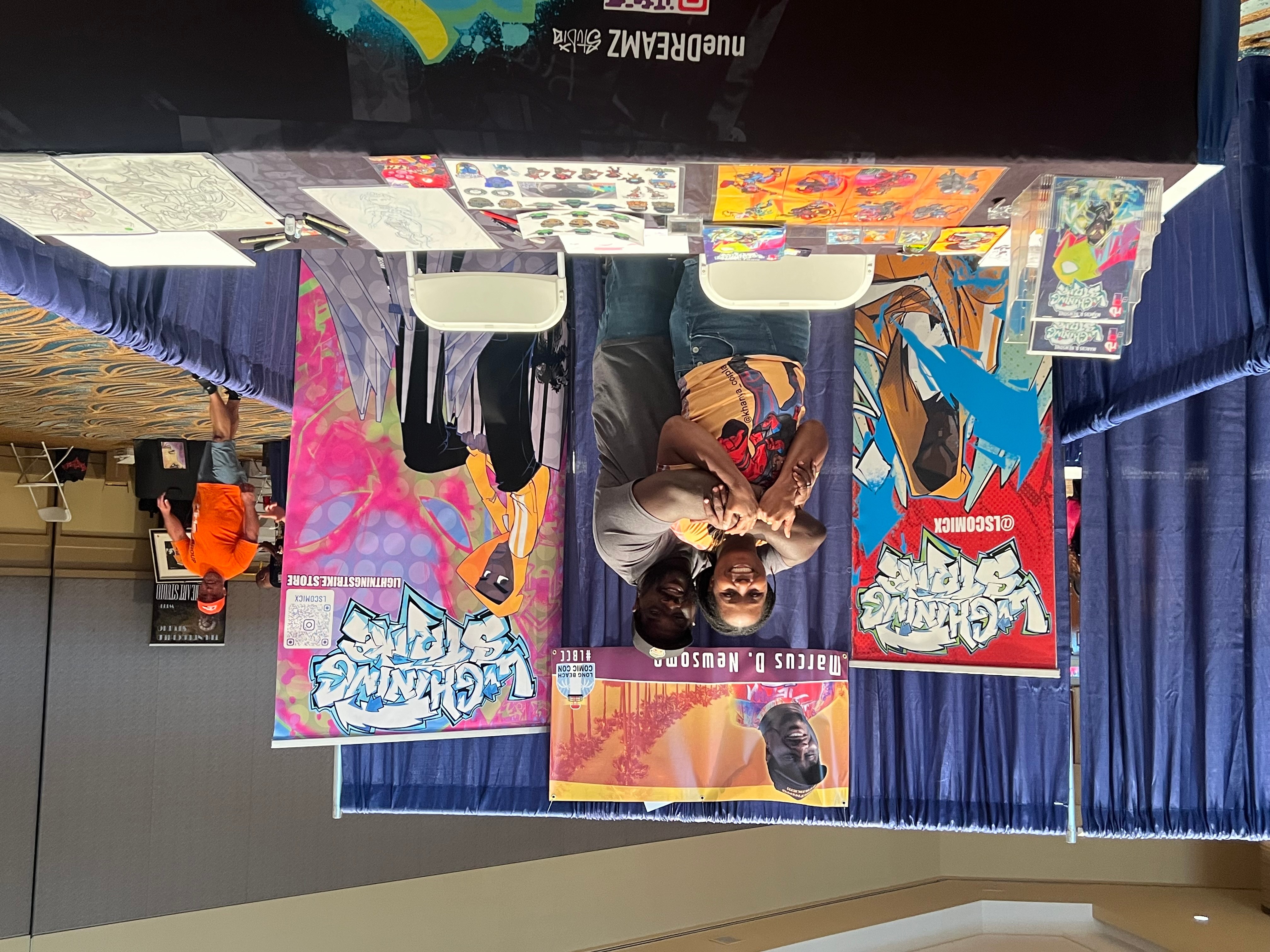 Table with colorful comic books and artwork, person sitting behind table, blue curtain backdrop, indoor setting.