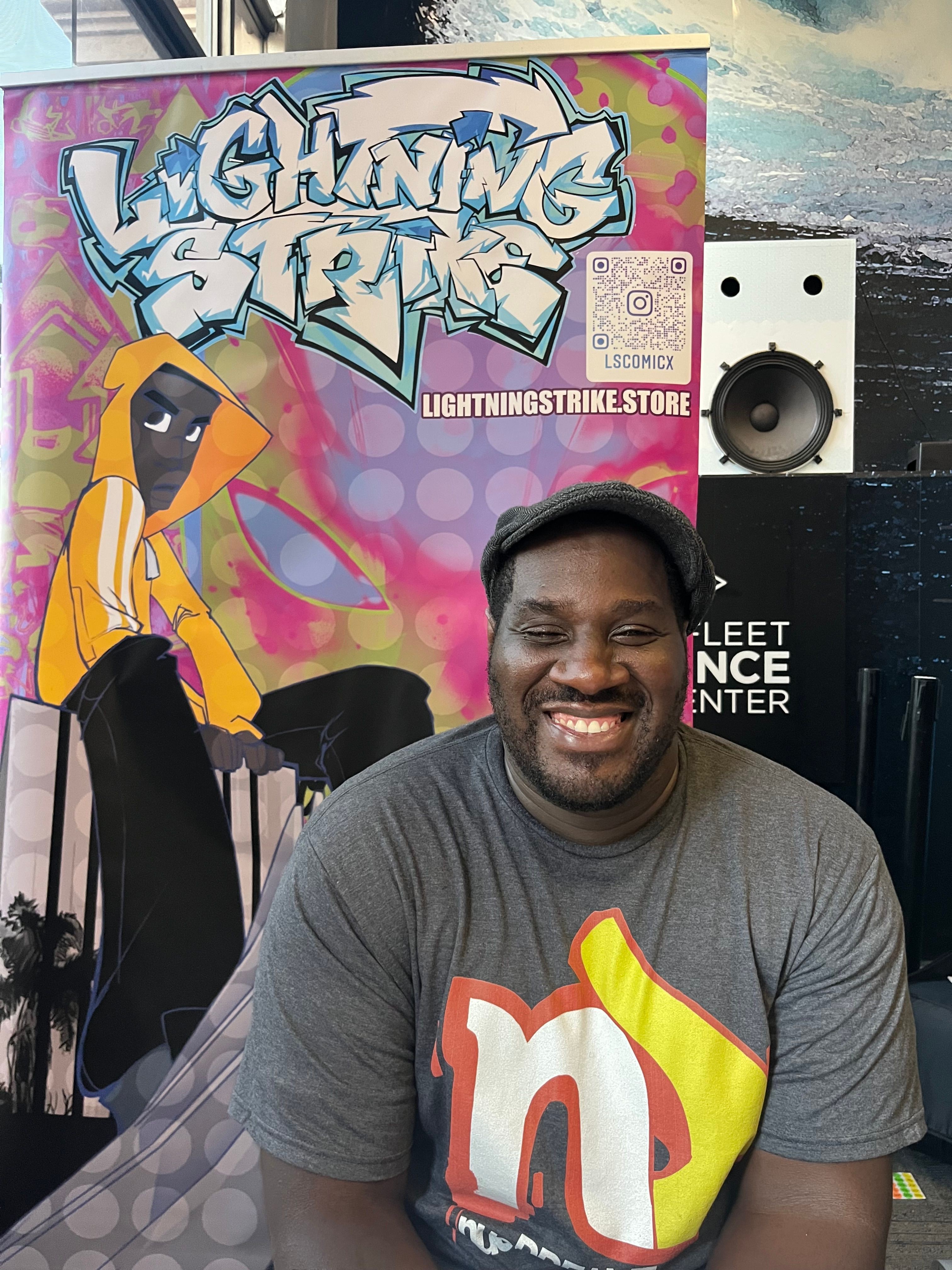 Man standing in front of graffiti-style artwork with colorful background, wearing a gray T-shirt with a red and yellow design.
