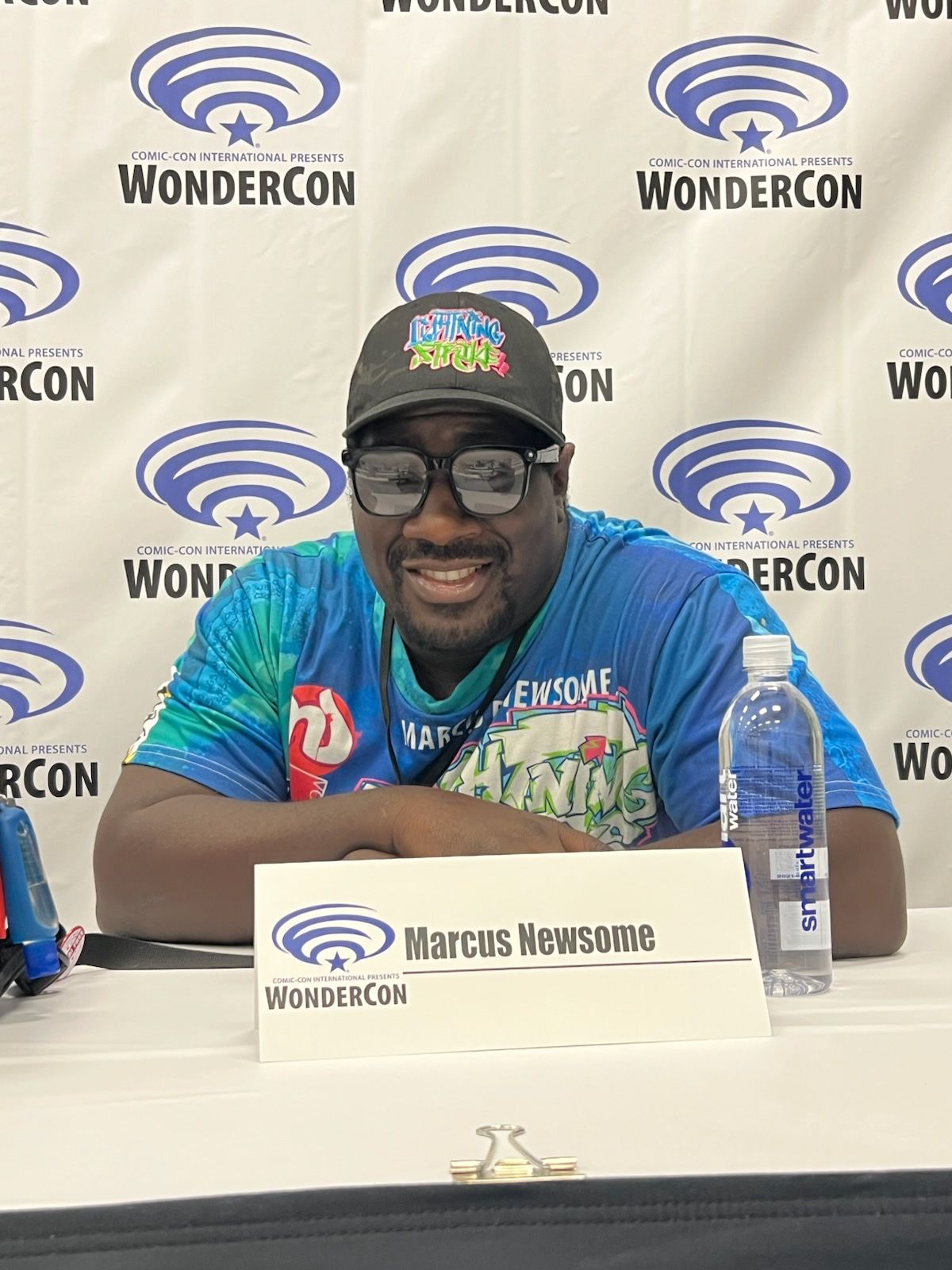 Person wearing a cap, glasses, and a colorful cycling jersey, sitting at a table with a water bottle and a sign, smiling.