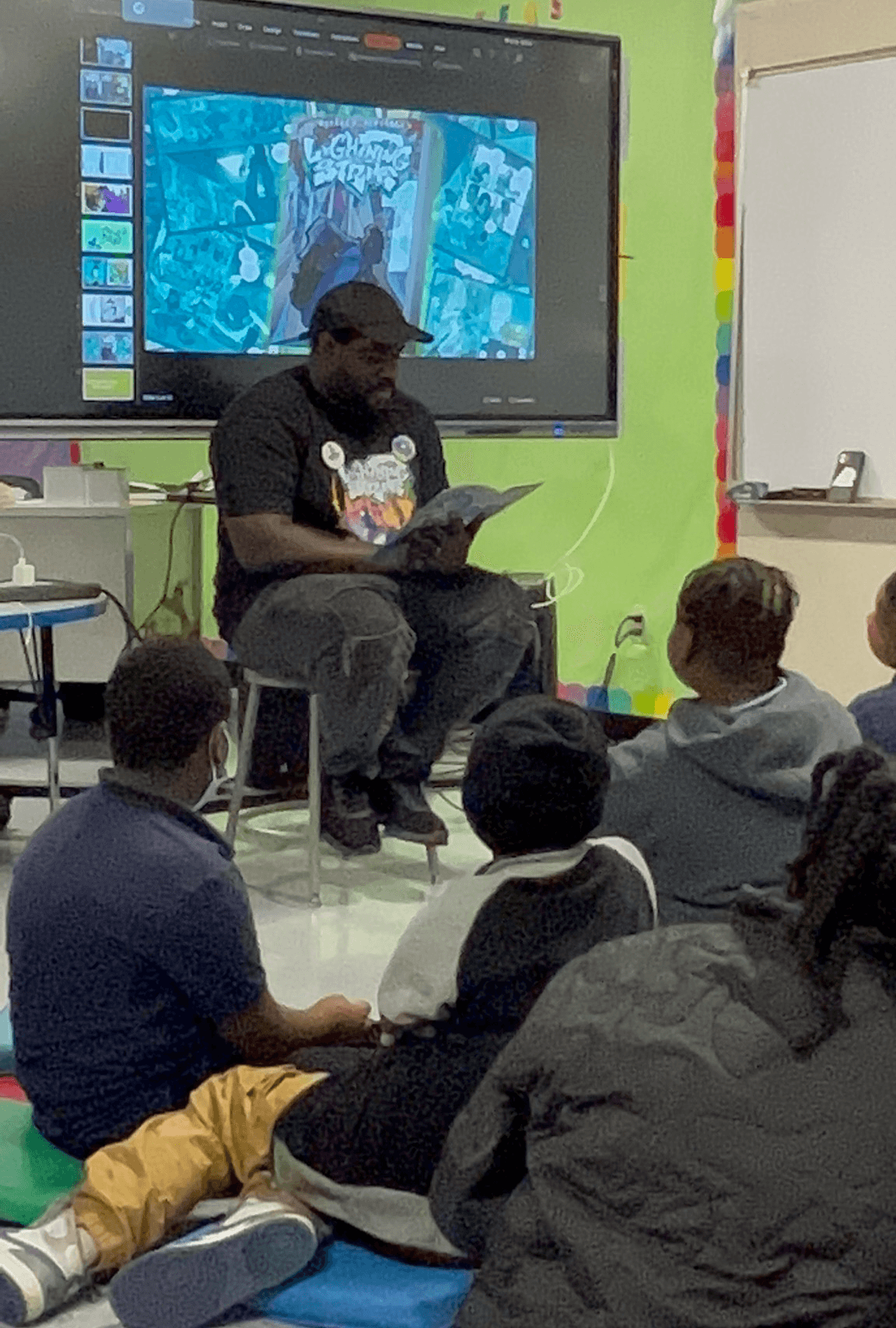 Person reading to children in a classroom with a large screen behind them displaying a colorful image.