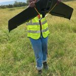 Person in a yellow safety vest holding a large black drone outdoors in a grassy field.