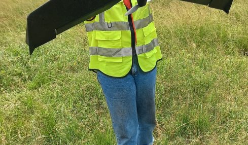 Person in a yellow safety vest holding a large black drone outdoors in a grassy field.