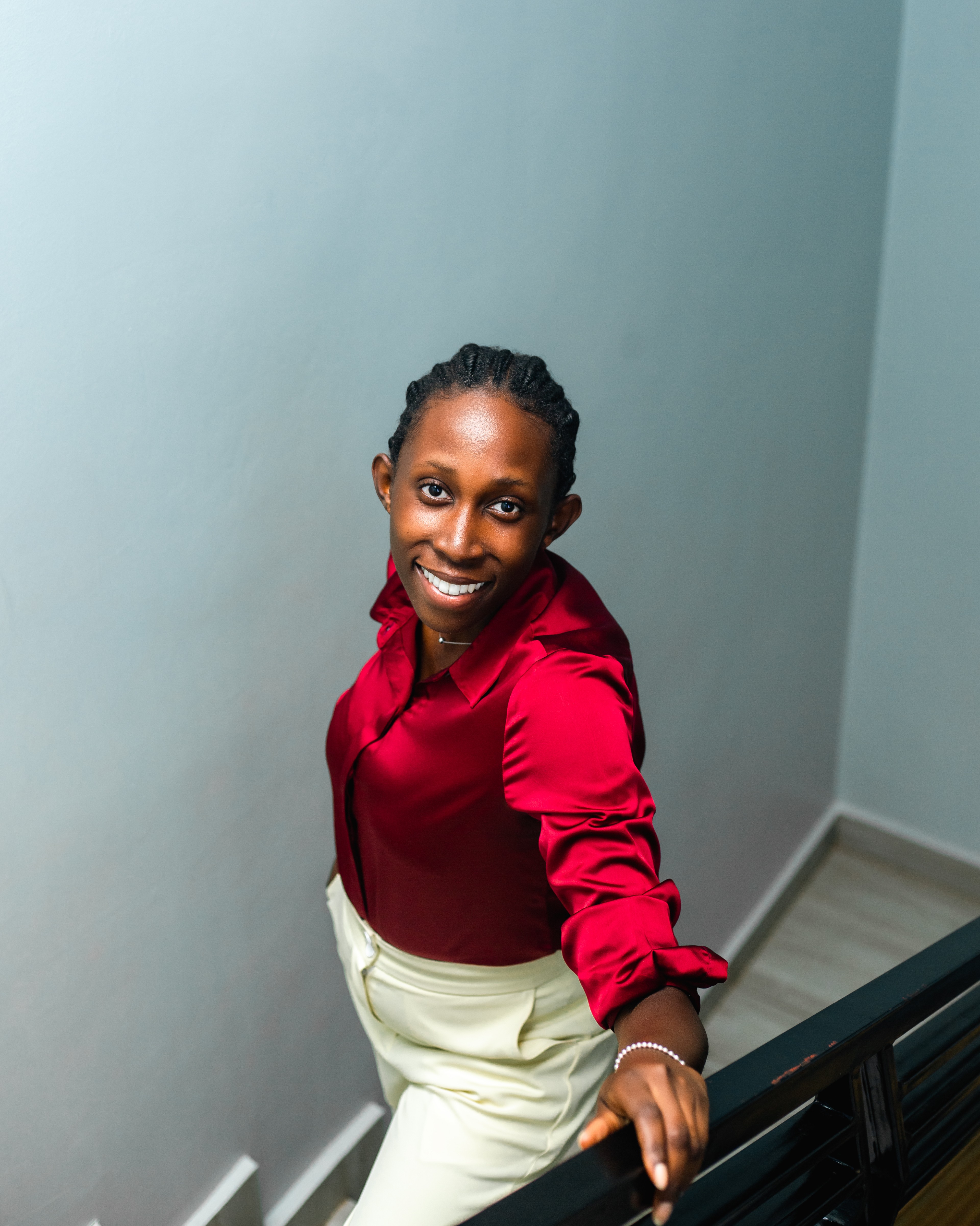 Smiling woman in red blouse and white pants standing near a staircase, looking up at the camera.