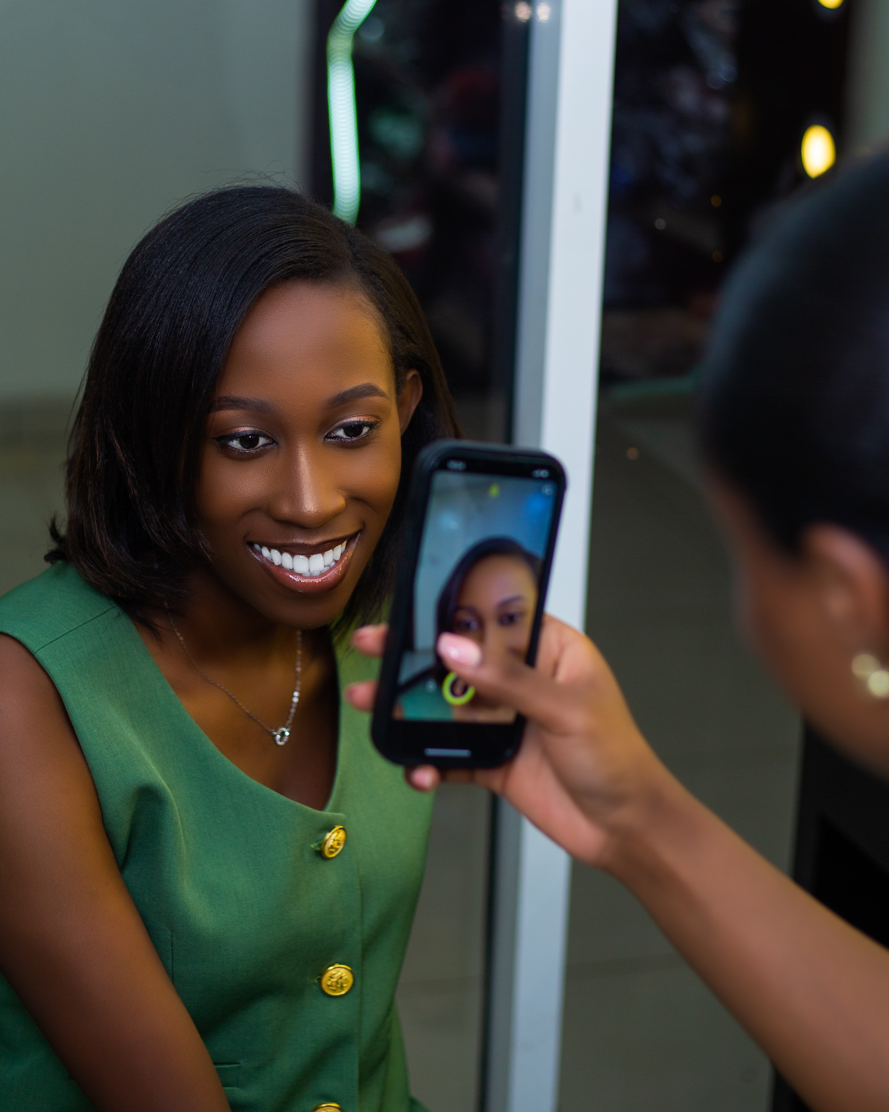 Woman taking a selfie with a smartphone, smiling, in an indoor setting with a mirror or glass behind her.