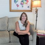 Woman sitting on a sofa in a living room, smiling, with a world map painting and a floor lamp behind her.