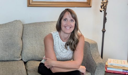 Woman sitting on a sofa in a living room, smiling, with a world map painting and a floor lamp behind her.