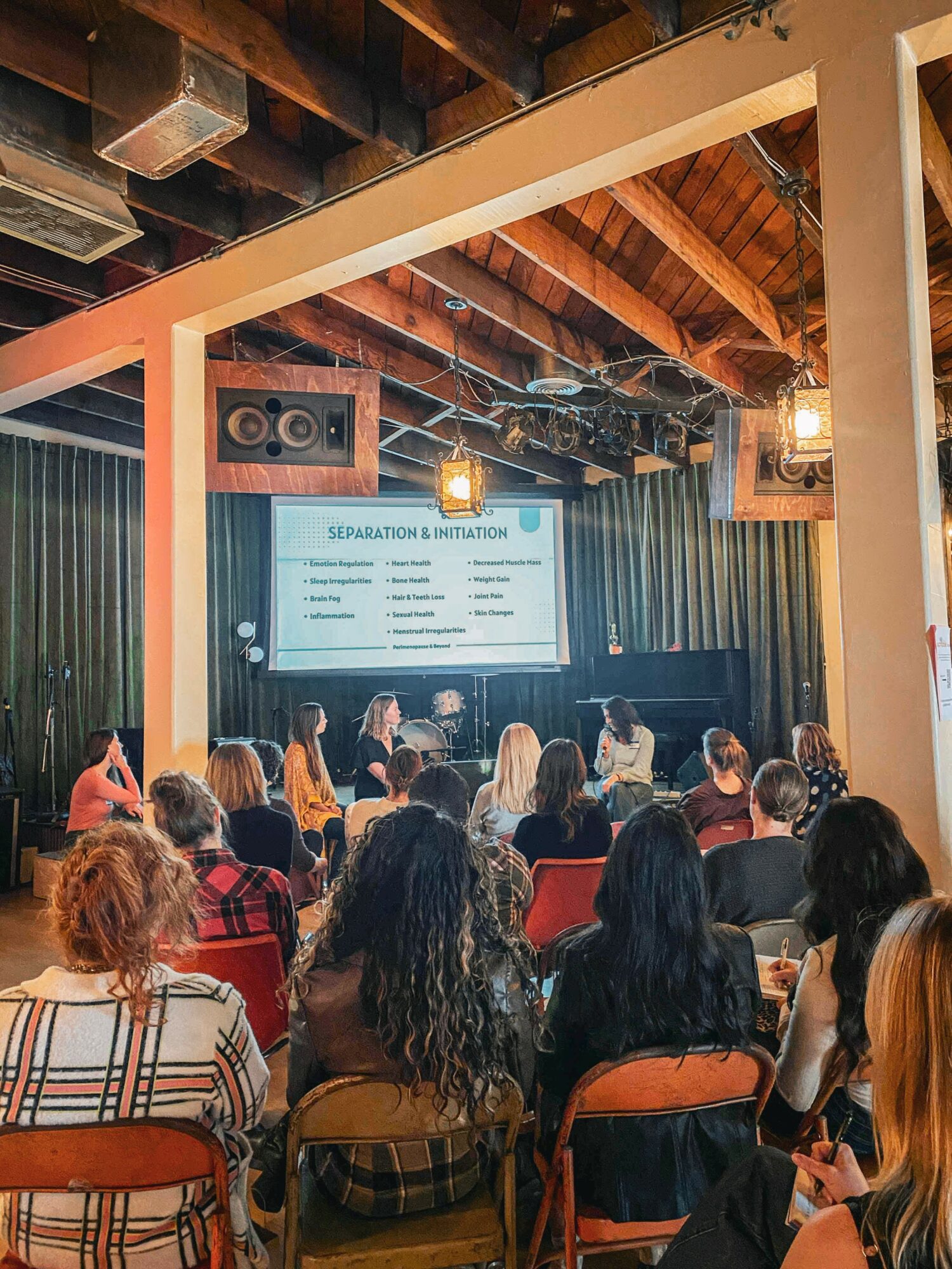 Audience watching a presentation in a room with wooden ceiling beams and a large screen displaying text.