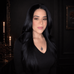 Woman with long black hair and a necklace, standing indoors with dark walls and candles in the background.