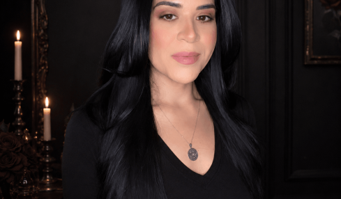 Woman with long black hair and a necklace, standing indoors with dark walls and candles in the background.