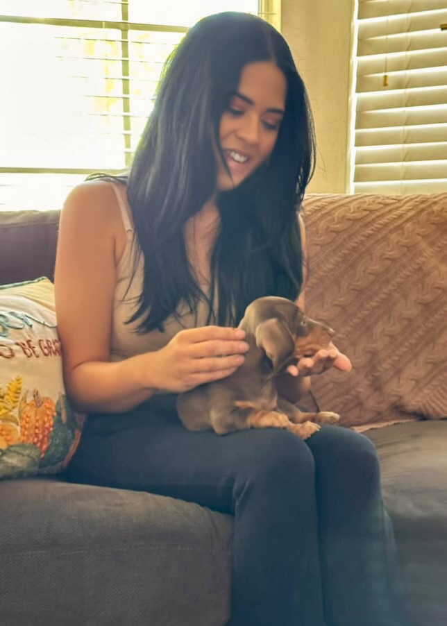 Woman with long dark hair sitting on a couch holding a small puppy, smiling, with window blinds behind her.