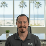 Man with dark hair and beard smiling, wearing a HLK managing shirt, sitting in an office with large window and beach view