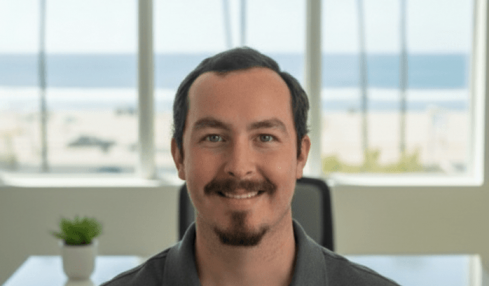 Man with dark hair and beard smiling, wearing a HLK managing shirt, sitting in an office with large window and beach view