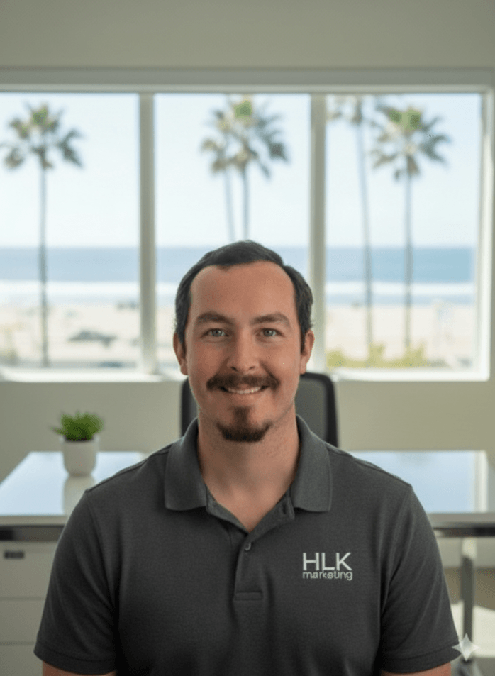 Man with dark hair and beard smiling, wearing a HLK managing shirt, sitting in an office with large window and beach view