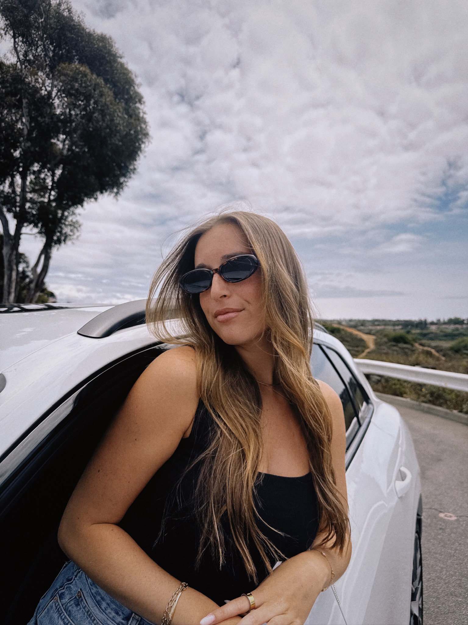 Woman with long hair wearing sunglasses and a black top leaning against a white car outdoors under cloudy sky.