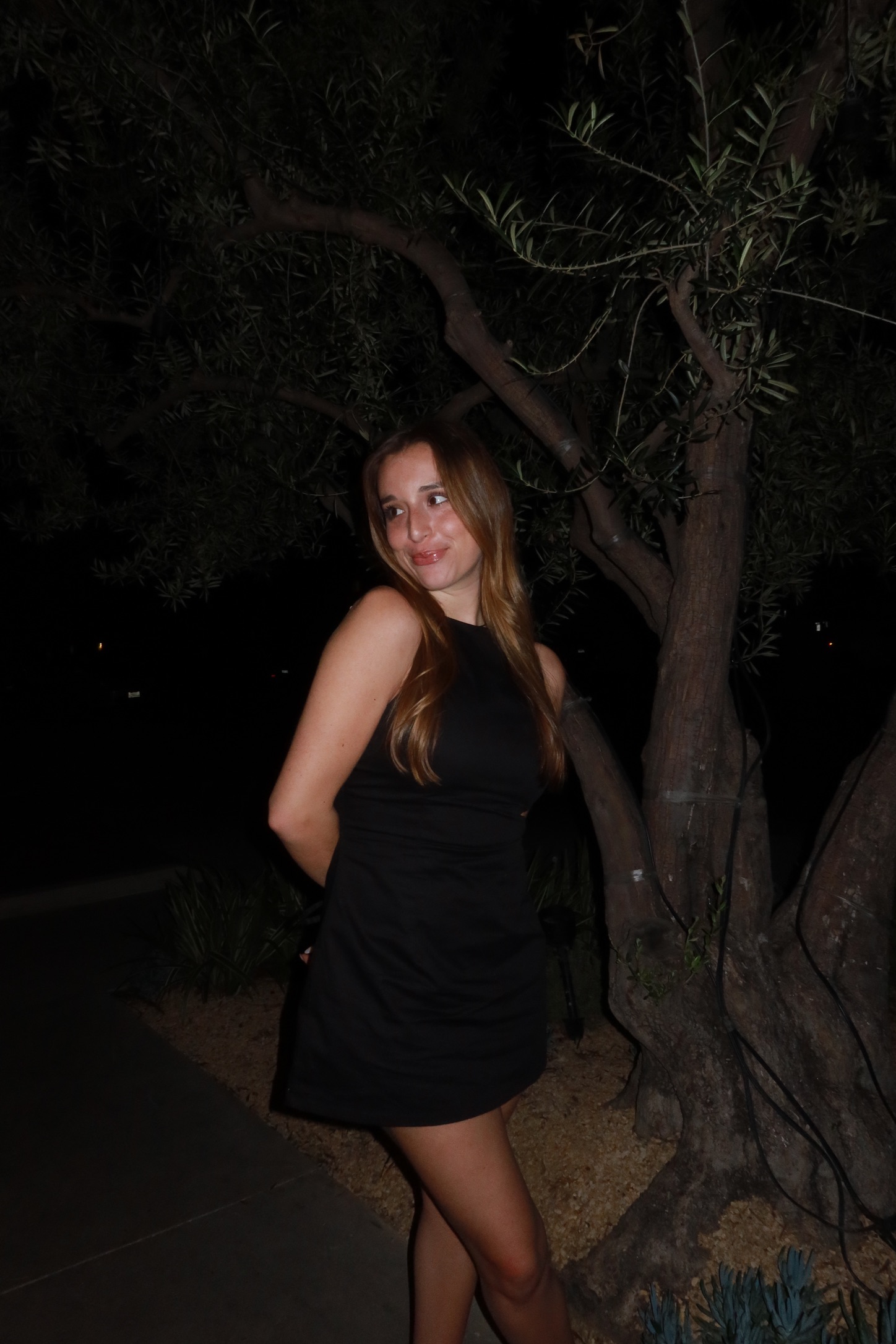 Young woman with long hair in a black dress standing outdoors at night near a tree.