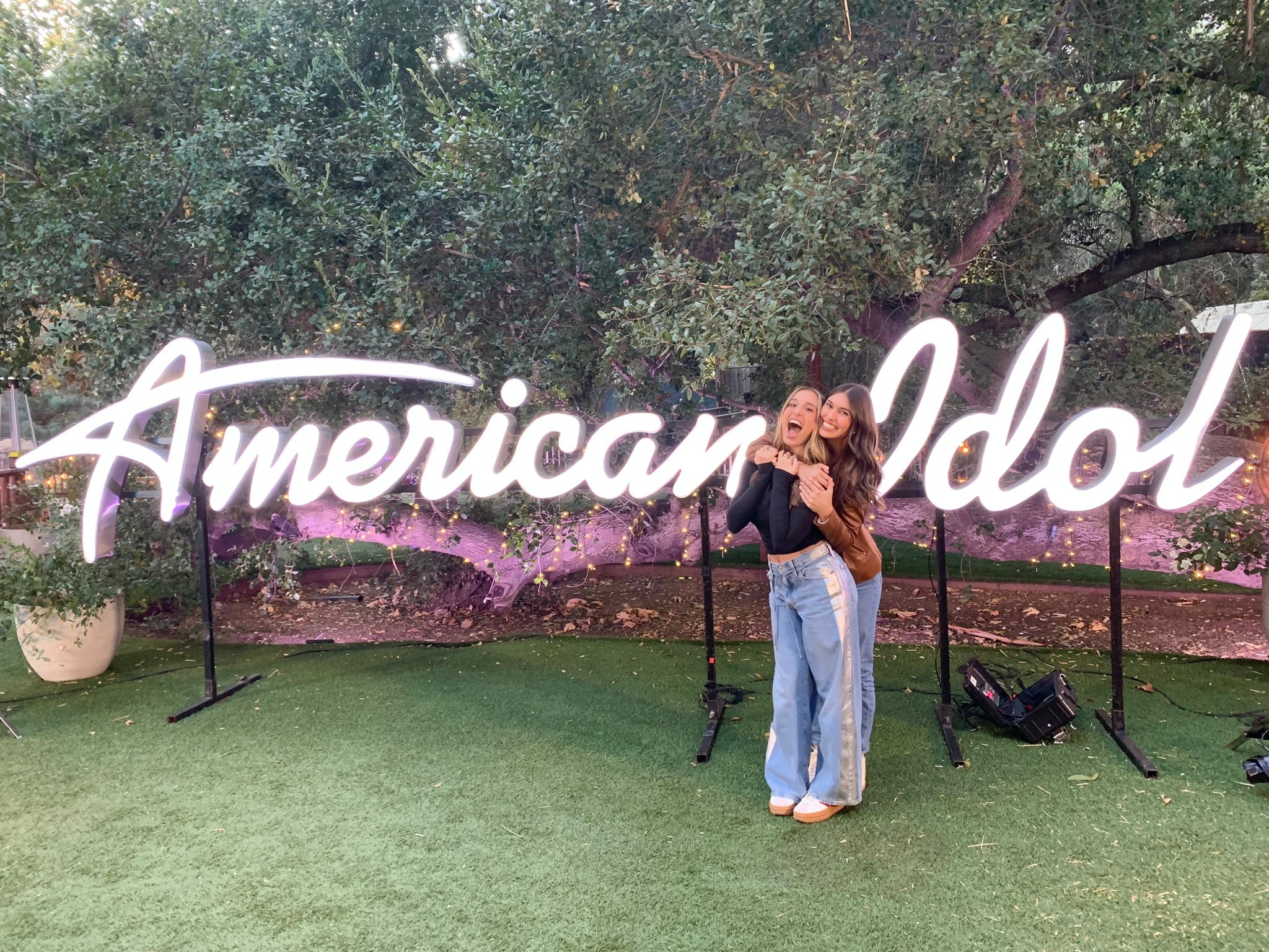 Two people hugging in front of a large illuminated 'American Idol' sign outdoors, with trees and grass in the background.