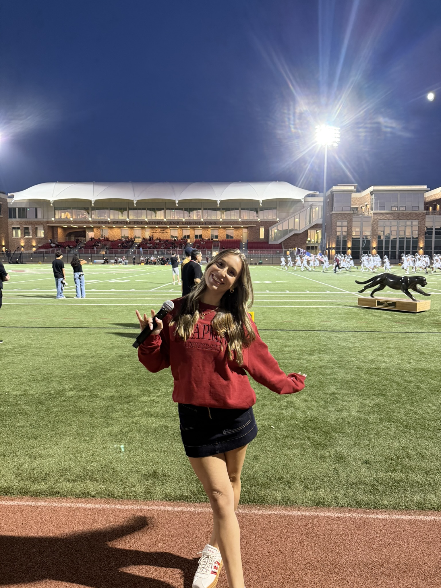 Young woman in red jacket and black skirt standing on a sports field at night, smiling, with stadium and bright lights behind.